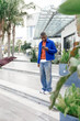 © Alina - A young African man stands on outdoor steps, wearing a blue jacket and orange shirt. He is looking at his smartphone amidst greenery and modern architecture.