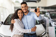 © Prostock-studio - Happy middle-eastern family buying new car in luxury showroom, standing buy brand new white auto, embracing and showing automatic key, copy space. Cheerful couple bought nice auto, car store interior