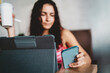 © BullRun - Focused woman holds phone while working on digital tablet, symbolizing tech-enabled independence, personal productivity and flexible control across devices.