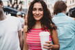 © BullRun - Young woman with coffee smiles directly at camera in busy city street, exuding warmth, approachability and personal charm in vibrant urban lifestyle.