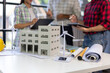 © Wasan - Miniature wind turbine and solar panel models are placed on a table alongside blueprints and a building model, while architects discuss a sustainable building project in the background.