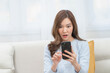 © Jestercine - Young Asian woman showing a shocked and surprised facial expression while looking at her smartphone, reacting to a scam message, fake news, or unexpected digital alert on a sofa at home.