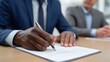© Bussakon - A business professional in a suit signs a legal document with a pen with a colleague in the background