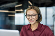 © Liubomir - Smiling woman wearing a headset and eyeglasses providing professional customer support service, having a friendly conversation during a video call in a modern office