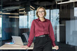 © Liubomir - Businesswoman with glasses and a professional maroon shirt standing confidently in a modern office space, leaning on a desk with a laptop, reflecting leadership and career success