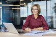 © Liubomir - Businesswoman with short blonde hair and glasses writing on documents and working on a laptop at a wooden desk, in a contemporary open-plan office setting