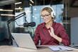 © Liubomir - Young woman smiling, wearing headphones and glasses, waving while recording a podcast or participating in a live online broadcast from her modern home studio setup with a microphone and laptop