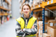 © Kowit - A young woman in a warehouse confidently poses with her arms crossed, wearing a high-visibility vest and a plaid shirt, surrounded by shelves of boxes.