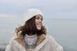 © SHOTPRIME STUDIO - Woman in a warm winter coat and beanie smiles softly outdoors by the sea, capturing a calm coastal moment with a gentle breeze and pale sky.