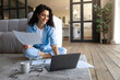 © Prostock-studio - Happy young Cauasian lady working or studying on laptop computer, holding documents, looking at pc screen indoors, empty space. Millennial freelancer doing research, having video conference at home
