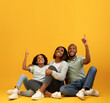 © Prostock-studio - Look there. Happy african american family pointing fingers up at empty space, sitting on floor over yellow background in studio. Free space for your advertisemet or text