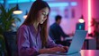 © miss irine - Young woman focused, typing on laptop computer in modern office. Evening work, neon lights glow, plants add nature. Another person works in background.