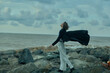 © SHOTPRIME STUDIO - Wind cloak sea rocks woman freedom in motion, dramatic landscape with breeze along coast, silhouette and energy meet horizon under changing weather, open air scene