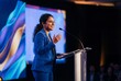 © Meow Creations - Confident indian woman in blue suit delivering a speech at a conference podium with microphone