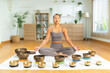 © Leny Studio - Meditation practitioner seated cross legged on mat with singing bowl set arranged for sound healing session in bright minimalist living room, calm focused expression