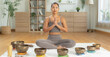 © Leny Studio - Young woman practicing seated meditation with hands in prayer pose among singing bowls on yoga mat in bright minimalist living room with plants and wooden furniture