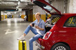 © Sofiia Shunkina - A woman sits in the open trunk of a red car, using her phone and enjoying a coffee break in a parking garage