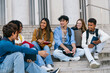 © Koldo_Studio - Group of diverse university students smiling and chatting happily while sitting on campus stairs outside the building