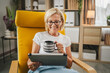 © Miljan Živković - Mature senior woman sit at home hold cup of hot drink and use tablet