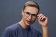 © SHOTPRIME STUDIO - Confident young man with glasses adjusting them, wearing casual blue sweater, looking directly at camera, poised and professional studio portrait on plain gray background, indoor setting, natural