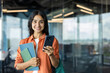 © Tetiana - Portrait of a young Indian woman standing indoors, holding books and a mobile phone, looking at the camera