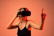 © Volodymyr_sh - Young woman using virtual reality headset and pointing forward in immersive digital environment with futuristic red lighting