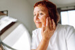 © maxbelchenko - Portrait of adult woman looking into small mirror and touching face while sitting in cozy room. Woman examines wrinkles, enjoys natural beauty indoors on sunny day. Concepts of aging, natural beauty.