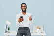 © SHOTPRIME STUDIO - Confident smiling man in white shirt holding mobile phone and giving thumbs up while standing at modern desk with laptop and stationery against light blue background, studio shot, lifestyle concept