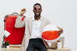 © SHOTPRIME STUDIO - Happy young man with dark hair and beard wearing sunglasses, white shirt, and beige blazer holding a beach ball, standing next to a large red suitcase in a bright studio setting. People lifestyle