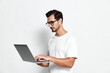 © SHOTPRIME STUDIO - Full body focused thoughtful young man wearing a white t-shirt using a laptop while standing on a solid white background. Technology work concept.