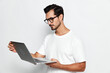 © SHOTPRIME STUDIO - Thoughtful man in stylish glasses holding laptop, looking at screen with concentration. Isolated on plain solid background, capturing focus and curiosity.