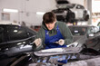 © JackF - Skilled young auto body technician in garage, wearing blue overalls and green hoodie, applying filler to damaged car bumper, preparing for painting..