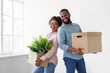 © Prostock-studio - Smiling young african american husband and wife holding cardboard boxes with plant in pot, on white wall background. Sweet home, moving to new apartment, married couple is relocation in own flat