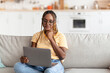© Prostock-studio - Attractive millennial african american woman with braids sitting on sofa, using modern notebook at home, unemployed young lady looking for job online, wearing eyeglasses, copy space