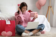 © Pixel-Shot - Beautiful young woman with laptop, gifts and shopping bags video chatting in bedroom. Valentine's Day celebration