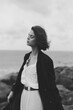 © SHOTPRIME STUDIO - Moody coastal scene with a woman in a dress and coat, wind-swept hair, standing along the shore, monochrome tones, elegant silhouette and calm atmosphere