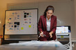 © Tj - Asian businesswoman working late in an office, holding a calculator and pen while reviewing financial reports and data on charts displayed on a whiteboard and computer screen