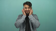 © Krakenimages.com - Young hispanic man closing eyes and covering ears over isolated green background wearing casual shirt, displaying frustration emotion, teal backdrop highlighting expression.
