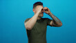 © Krakenimages.com - Young man with tattoos makes binocular hands gesture against vibrant blue background showing playfulness and curiosity in an expressive way.