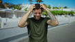 © Krakenimages.com - Hispanic man with tattoos stands outdoor in an urban street setting, appearing worried with hands on his head, wearing a green shirt and displaying expressive emotions.
