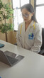 © Krakenimages.com - Young chinese woman doctor wearing uniform with stethoscope, focused on a laptop in a bright clinic room, emphasizing a professional healthcare environment.