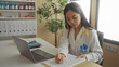 © Krakenimages.com - Young chinese woman doctor with stethoscope in hospital clinic room working on laptop and reviewing documents.