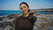 © Krakenimages.com - Young hispanic woman outdoors at seaside beach smiling with arm extended towards camera under clear sky