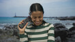 © Krakenimages.com - Woman smiling holding controller against her ear by seaside with rocks and ocean, wearing striped shirt, young hispanic enjoying outdoors on beach day.