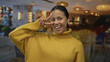 © Krakenimages.com - Young hispanic woman in yellow sweater makes playful peace sign inside a warmly lit hotel indoors, showcasing a joyful and friendly atmosphere.