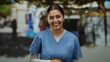 © Krakenimages.com - Hispanic woman nurse smiling outdoors, wearing blue scrubs, set against a blurred urban street with trees, conveying warmth and professionalism in a public space.
