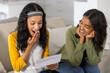 © wavebreak3 - Diverse mom and teenage daughter sitting on beige sofa in living room reading letter together