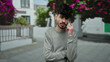 © Krakenimages.com - Young man with dark hair and beard in gray sweater talking on smartphone outside on urban street with flowers blurred in background