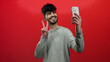 © Krakenimages.com - Young man taking selfie over red background using smartphone smiling confidently while showcasing peace sign and wearing casual gray sweater