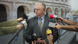 © Krakenimages.com - Senior man in suit with clasped hands and closed eyes facing multiple handheld microphones from reporters in front of an ancient colosseum on a street; concern.